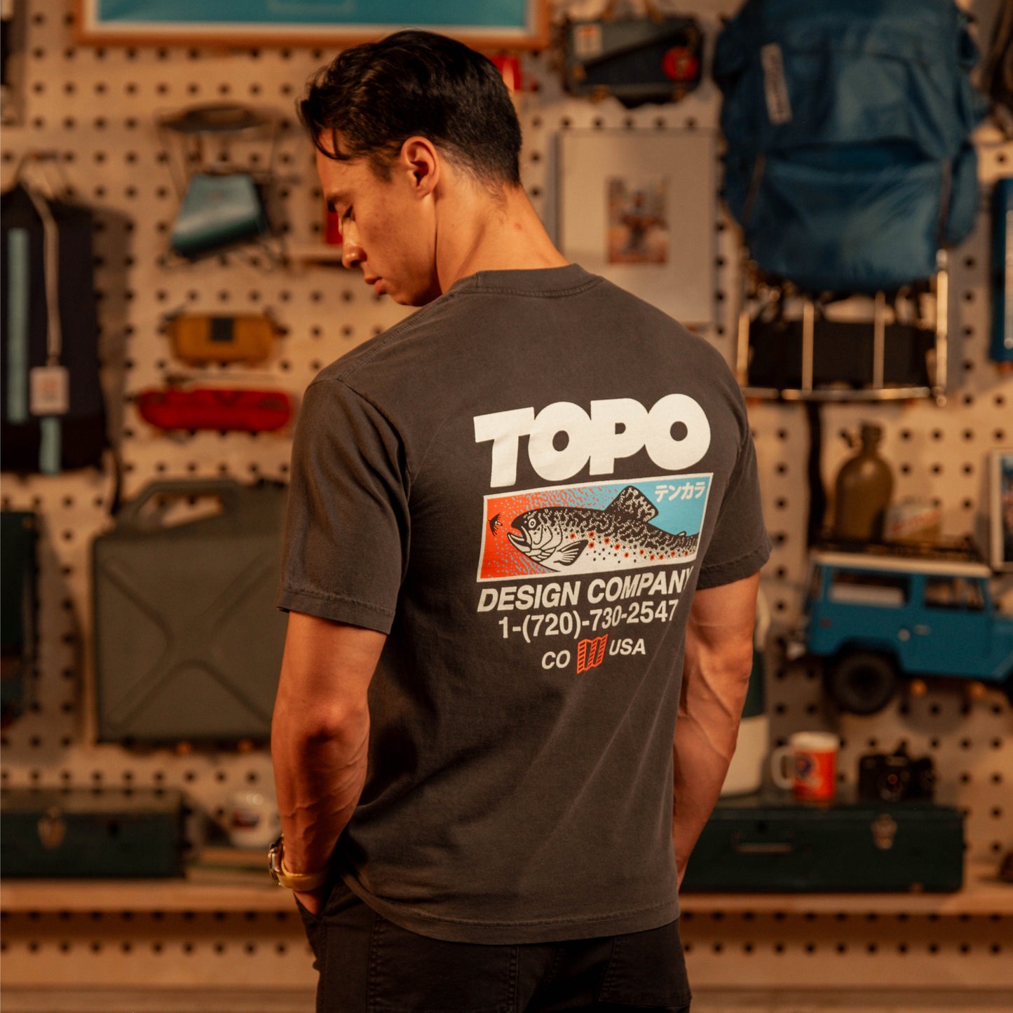Man wearing black graphic t-shirt facing away towards wall with camping gear and vintage bags
